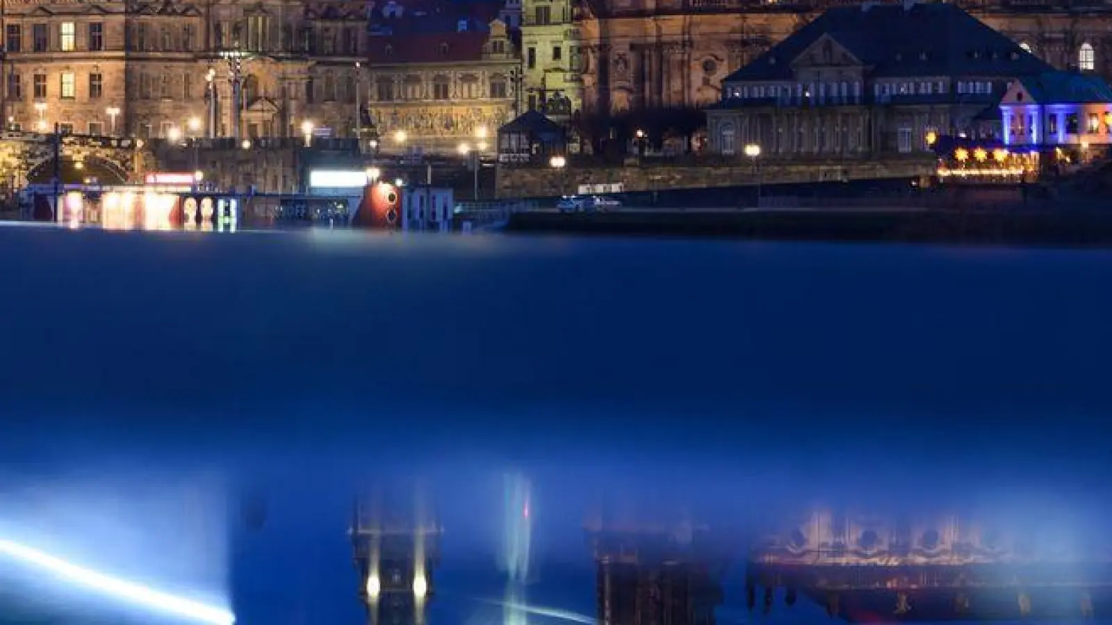 Scheinwerfer vorbeifahrender Radfahrer ziehen auf dem Elberadweg vor der Altstadtkulisse in Dresden Lichtspuren und spiegeln sich dabei in einer Pfütze. (Foto: Robert Michael/dpa)