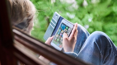 Ein Junge sitzt am Fenster und schaut auf sein Handy. (Foto: Annette Riedl/dpa/Symbolbild)