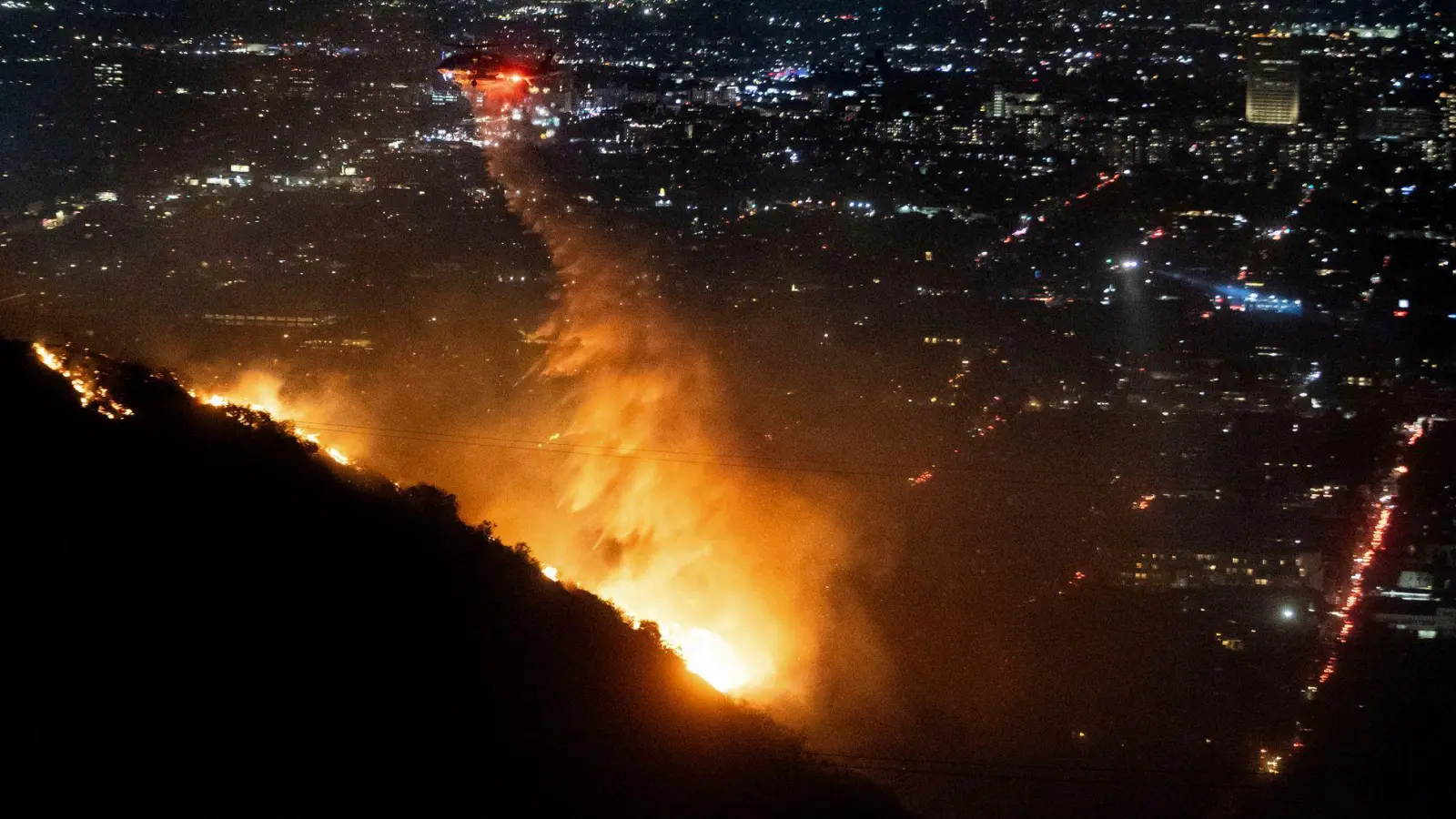 Ein Hubschrauber wirft Wasser auf das brennende Sunset Fire in den Hollywood Hills. (Foto: Ethan Swope/AP/dpa)