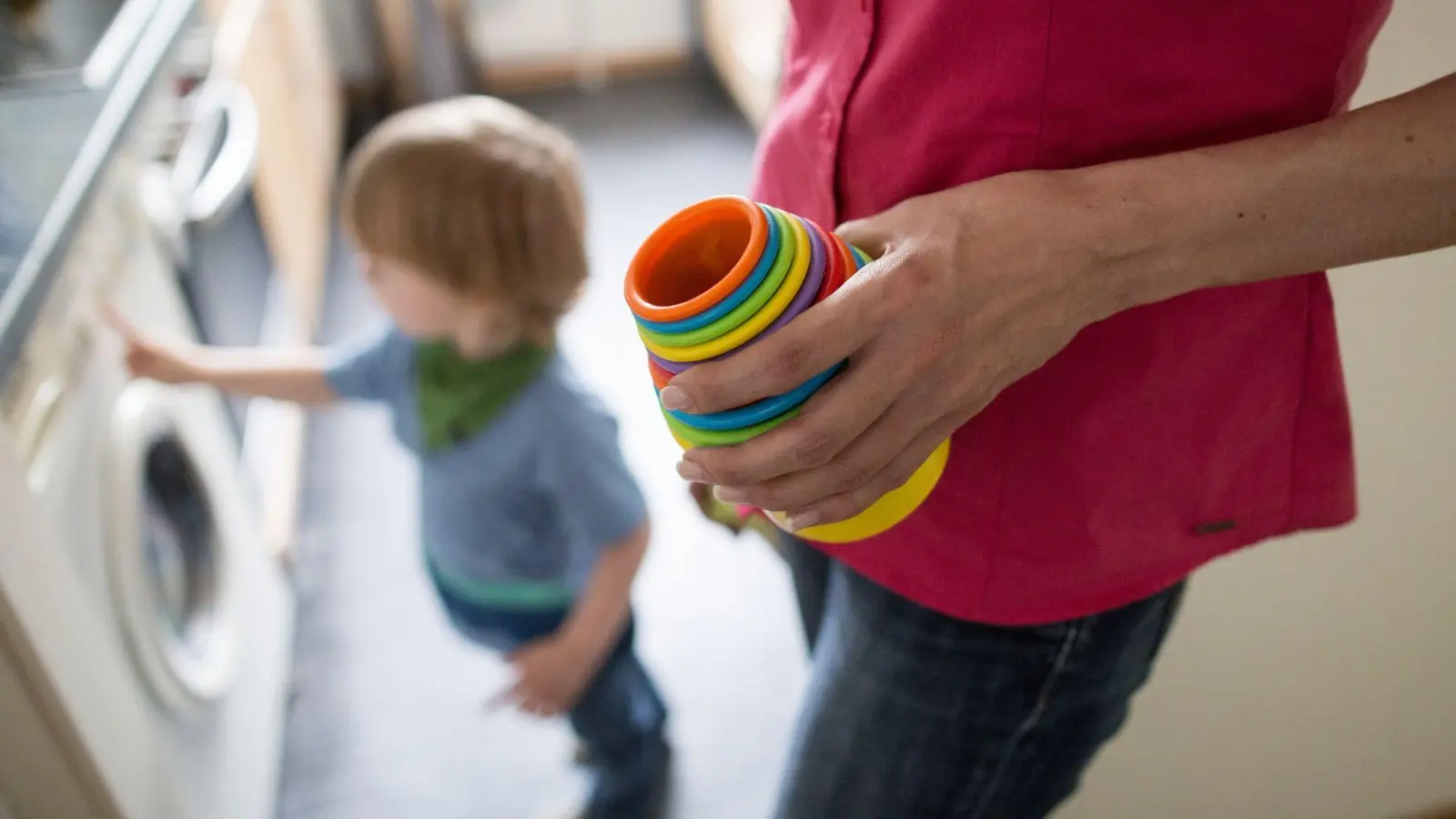 Eine Mutter hält Kinderspielzeug in der Hand, während ein kleiner Junge an einer Waschmaschine spielt. Die Krisen der letzten Jahre belasten vor allem Mütter - Stress und finanzielle Probleme sorgen für eine Dauerbelastung. (Foto: Marcel Kusch/dpa)