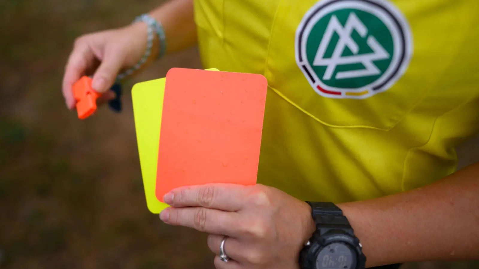 Eine Schiedsrichterin, steht mit roter und gelber Karte auf einem Rasen und hält in der anderen Hand eine Trillerpfeife. (Foto: Robert Michael/dpa/Archivbild)