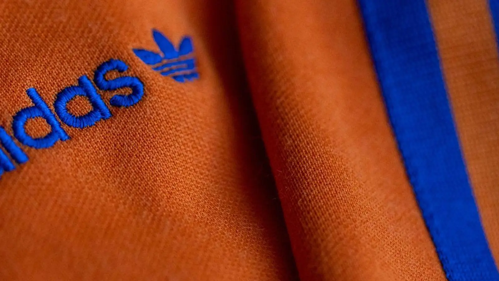 Das Logo des Sportartikelherstellers adidas in blau auf einer orangen Jacke. (Foto: Daniel Karmann/dpa)