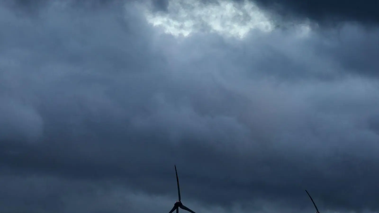 Windräder stehen bei Aitrang im Allgäu unter dichten Regenwolken hinter einem Wald. (Foto: Karl-Josef Hildenbrand/dpa/Archivbild)