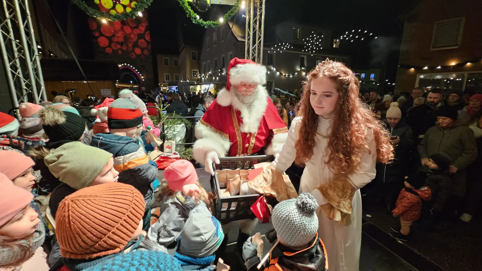 Bescherung für die kleinen Sängerinnen und Sänger aus dem Kindergarten „Regenbogen“: Christkind Emily Schwab und Weihnachtsmann Uwe Lechner hatten bei der Weihnachtsdorf-Eröffnung am Freitagabend in Leutershausen Geschenke dabei. Im Hintergrund das heuer erstmals bunt beleuchtete Obere Tor. (Foto: Wolfgang Grebenhof)