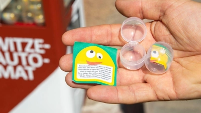 Der Kabarettist Oliver Tissot hält vor seinem Witze-Automaten Plastik-Kapseln, die mit kleinen Zetteln mit je einem Witz bestückt sind, in der Hand. (Foto: Daniel Karmann/dpa/Archivbild)