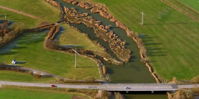 Das Foto zeigt die auf diesem von oben nach unten fließende renaturierte beziehungsweise umgestaltete Altmühl bei Ornbau mit der Staatsstraße 2411 Triesdorf-Arberg. Auf dem rechten Flussufer oben ist der Schilfgürtel zu sehen, der sich so stark ausgebreitet hat, dass etwa Fischern dort der Zugang zum Wasser stark erschwert wird. Das Schilf wird durch eine Beweidung mit Angus-Rindern eingedämmt. (Foto: Hajo Dietz)