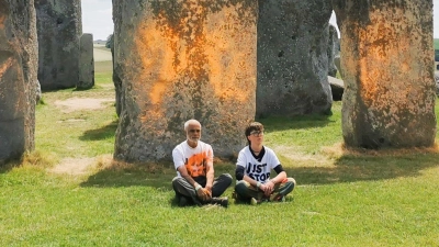 Demonstranten von „Just Stop Oil“ sitzen vor dem Steinmonument Stonehenge, das sie zuvor mit orangener Farbe besprüht haben. (Foto: Just Stop Oil/PA Media/dpa)